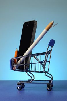 A miniature shopping cart with a cell phone and writing tools inside over a blue background.