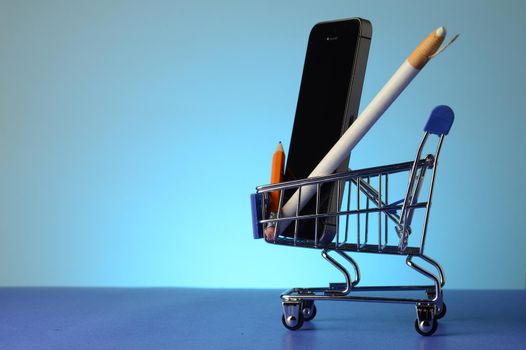 A miniature shopping cart with a cell phone and writing tools inside over a blue background.