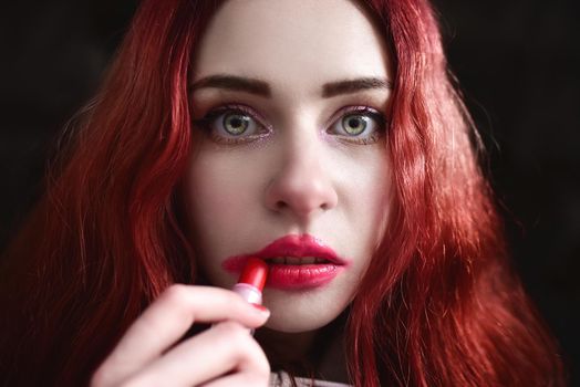 Portrait of crazy-looking depressed teen girl with red hair she is smearing red lipstick on her face, black background. social issues. social protest. The problem with raising teenagers