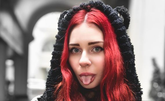Close portrait of a smiling teen girl with red hair in warm clothes standing outside and looks into the camera and jokely showing her tongue.