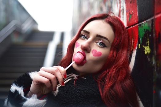 A glamorous sexy teenage girl with red hair licks a sweet strawberry candy near a wall of graffiti.