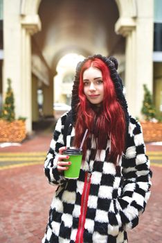 Near portrait of a teen girl with red hair in warm clothes standing outside on a cold day with a cup of coffee in her hands and looks into the camera. Cute girl walking with coffee in her hands.