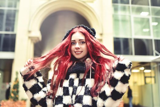 Near portrait of a smiling teen girl with red hair in warm clothes standing outside and looks into the camera and playing with own hair.