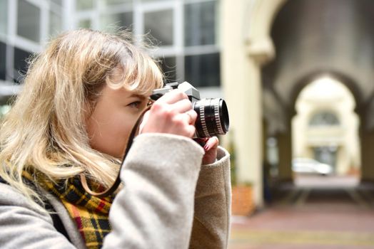 Blonde pretty photographer woman with retro camera in her hands while taking pictures at urban old architecture. Discover new places.