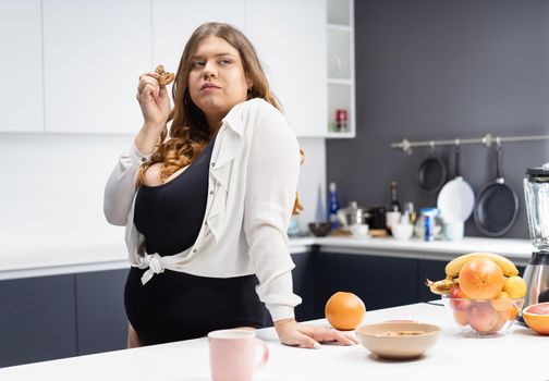 Lost battle eating cookie overweight girl standing at kitchen. Dieting and nutrition concept. Happy curvy body young woman with long blond hair at modern kitchen.