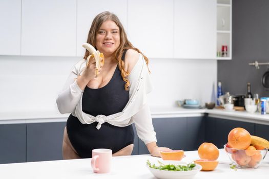 Dieting and nutrition concept. Happy curvy body young woman with long blond hair using blender at modern kitchen, blending fresh fruits for healthy smoothie.