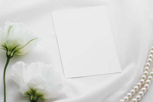 Wedding invitation, white rose flowers and pearls on silk fabric as bridal flatlay background, blank paper greeting card and holiday branding, flat lay design concept