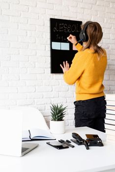 Distance learning. E-learning. Young smiling woman in yellow sweater and black headphones teaching online using video chat on laptop , writing on blackboard. Focus on foreground