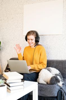 Distance learning. E-learning. Young smiling woman in yellow sweater and black headphones studying online using laptop, sitting on the couch at home