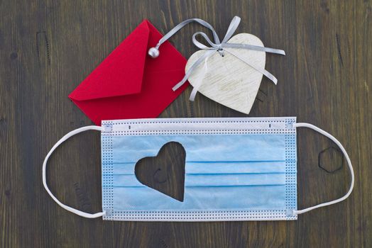 Face mask with cut out heart shape and red envelope on wooden background with free copy space. Social distancing and contact-less greetings
