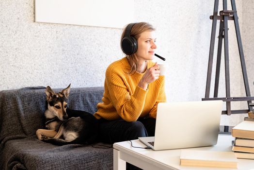 Distance learning. E-learning. Young smiling woman in yellow sweater and black headphones studying online using laptop, sitting on the couch at home, drinking coffee