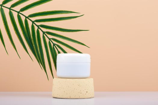 White cream jar face cream, mask or scrub on podium against beige background with palm leaf and copy space. Concept of skin care and beauty