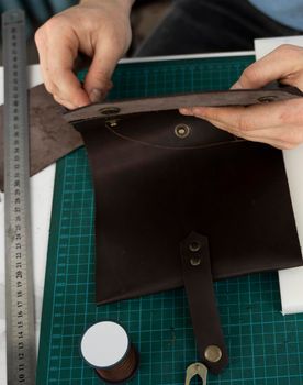 Mens hand working on the leather wallet in his workshop. Working process with a brown natural leather. Craftsman holding a crafting tools