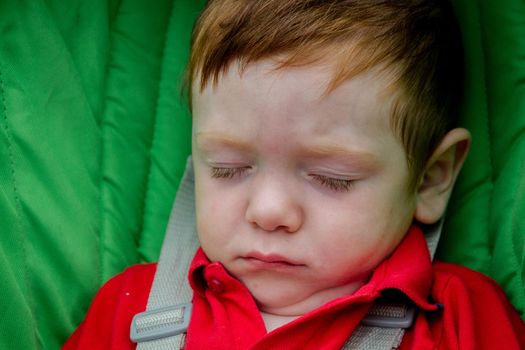 Portrait of a cute, redhead baby boy wearing a red shirt and sleeping in a green pushchair