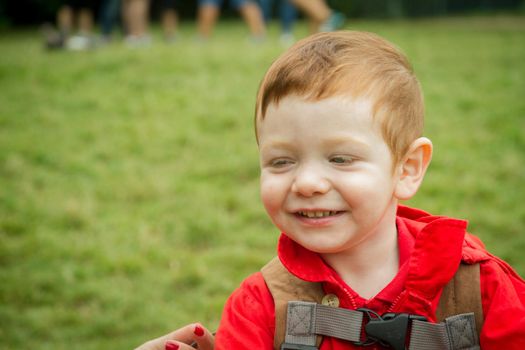 Portrait of a cute, blue-eyed, redhead baby boy smiling wearing a red jacket in a park.