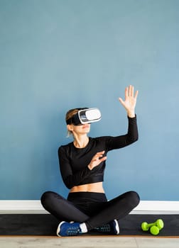 Fitness, sport and technology. Young athletic woman wearing virtual reality glasses sitting at fitness mat using VR interactive menu at blue background