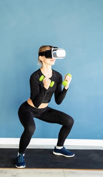 Fitness, sport and technology. Young athletic woman wearing virtual reality glasses squatting with dumbbells at blue background