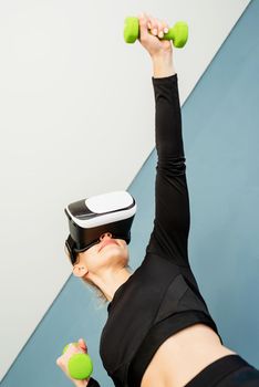 Fitness, sport and technology. Young athletic woman wearing virtual reality glasses standing at fitness mat working out with dubbells