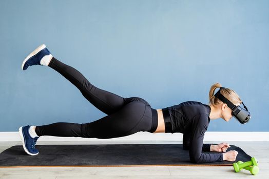 Fitness, sport and technology. Young athletic woman wearing virtual reality glasses doingplank at blue background