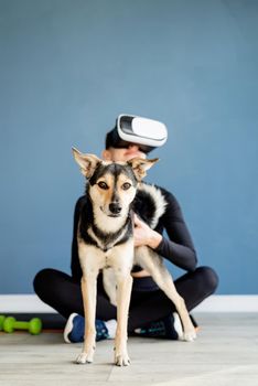 Fitness, sport and technology. Young athletic woman wearing virtual reality glasses sitting on fitness mat with dog