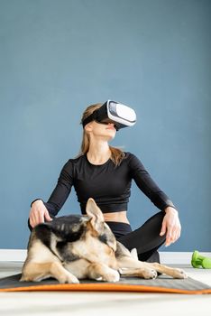 Fitness, sport and technology. Young athletic woman wearing virtual reality glasses sitting on fitness mat with dog