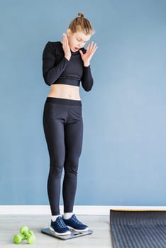 Dieting and weight loss. Young surprised woman in black sport clothes standing on scales