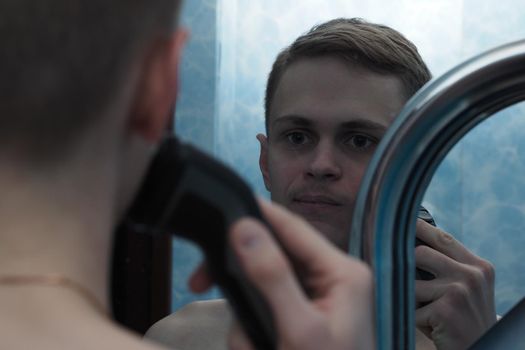 A young man of European appearance shaves his face with an electric razor and looks in the mirror. High quality photo