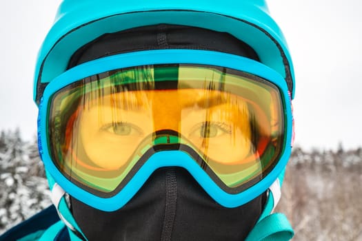 Female in ski mask with blue eyes. Snowboarder looking at camera. Behind there is a beautiful view of the snow-covered forest. Ski vacation. Logoisk, Belarus