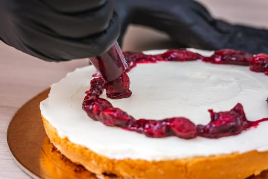 Female hand in black glove squeezes out cherry confiture on a cheesecake with coconut.