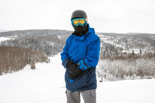 Male in a blue sports suit standing a large snow slope. Snowboarder looking at a camera. Winter forest in the background. Skiing holidays. Close-up. Logoisk, Belarus