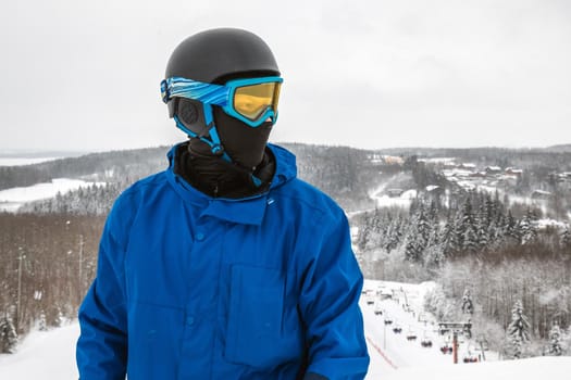 Male in a blue sports suit standing a large snow slope. Snowboarder Snowboarder looks at a beautiful view. Winter forest in the background. Skiing holidays. Close-up. Logoisk, Belarus