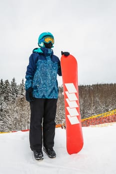 Female in a blue suit standing with a snowboard on a large snow slope. Snowboarder looks at a beautiful view of the snowy forest. Skiing holidays. Logoisk, Belarus