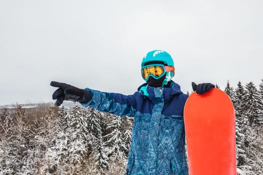 Female in a blue suit standing with a snowboard on a large snow slope. Snowboarder looks at a beautiful view of the snowy forest. Skiing holidays. Logoisk, Belarus