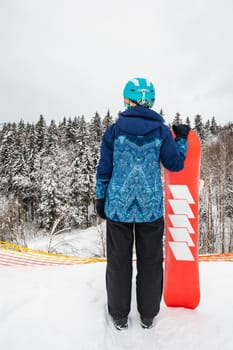 Female in a blue suit standing with a snowboard on a large snow slope. Snowboarder looks at a beautiful view of the snowy forest. Skiing holidays. Logoisk, Belarus