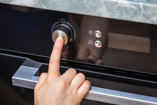 Female hand turning on the oven to make a cake at home. Close-up of the oven knob. View of the black electric oven, knob with a hot air icon. The concept of heating food, baking a cake.
