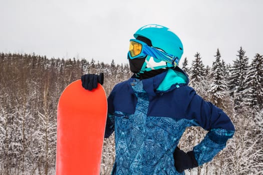 Female in a blue suit standing with a snowboard on a large snow slope. Snowboarder looks at a beautiful view of the snowy forest. Skiing holidays. Logoisk, Belarus