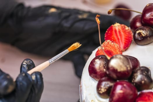 Female hand in black glove puts edible gold on cherries and strawberries on top of cheesecake with coconut.
