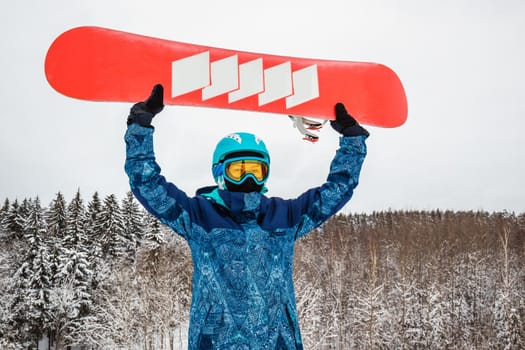 Female in a blue suit standing with a snowboard on a large snow slope. Snowboarder holds the snowboard over his head. Skiing holidays. Logoisk, Belarus