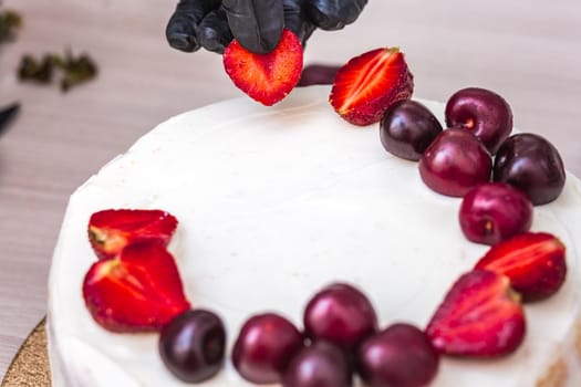 Female hand in black glove puts cherries and strawberries on top of cheesecake with coconut.
