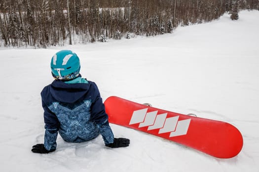 Female in a blue suit sitting with a snowboard on a large snow slope. Snowboarder looks at a beautiful view of the snowy forest. Skiing holidays. Logoisk, Belarus