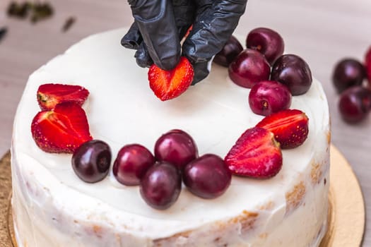 Female hand in black glove puts cherries and strawberries on top of cheesecake with coconut.