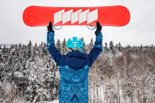 Female in a blue suit standing with a snowboard on a large snow slope. Snowboarder holds the snowboard over his head. Skiing holidays. Logoisk, Belarus