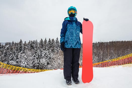 Female in a blue suit standing with a snowboard on a large snow slope. Snowboarder looks at a beautiful view of the snowy forest. Skiing holidays. Logoisk, Belarus