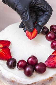 Female hand in black glove puts cherries and strawberries on top of cheesecake with coconut.