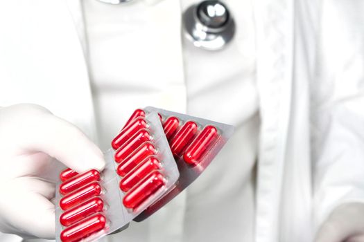 Doctor in white surgical gloves and wearing stethoscope holding red capsule blisters