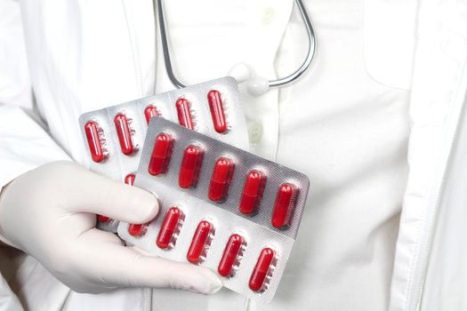 Doctor in white surgical gloves and wearing stethoscope holding red capsule blisters