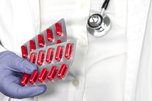 Doctor in blue surgical gloves and wearing stethoscope holding red capsule blisters