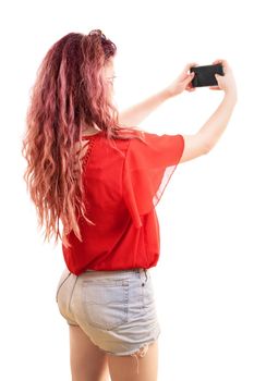 Back/ side view of a beautiful fashionable redhead young woman taking a selfie with a smartphone, isolated on a white background. Copy space.