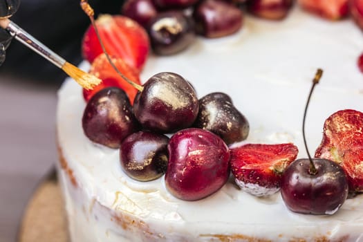 Female hand in black glove puts edible gold on cherries and strawberries on top of cheesecake with coconut.