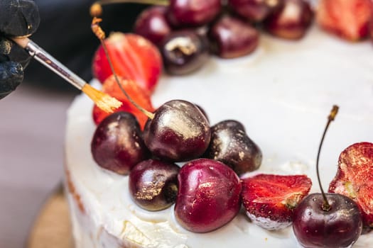 Female hand in black glove puts edible gold on cherries and strawberries on top of cheesecake with coconut.
